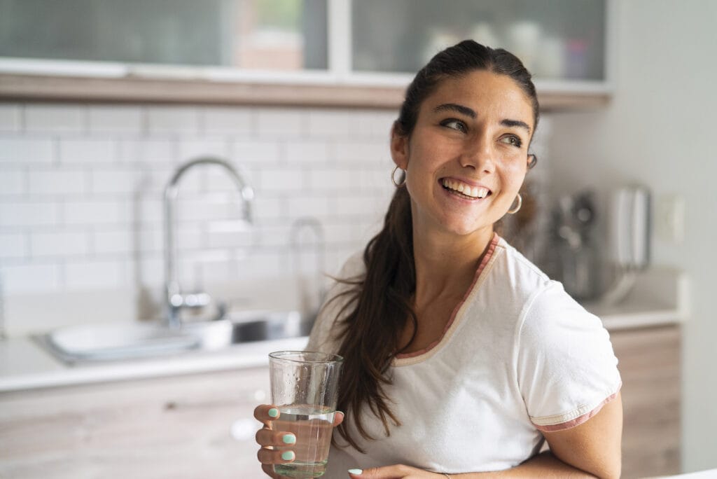 Benefits of a plumbing maintenance plan. Front shot of beautiful smiling young adult hispanic woman holding glass of water while looking up standing in kitchen during daytime.